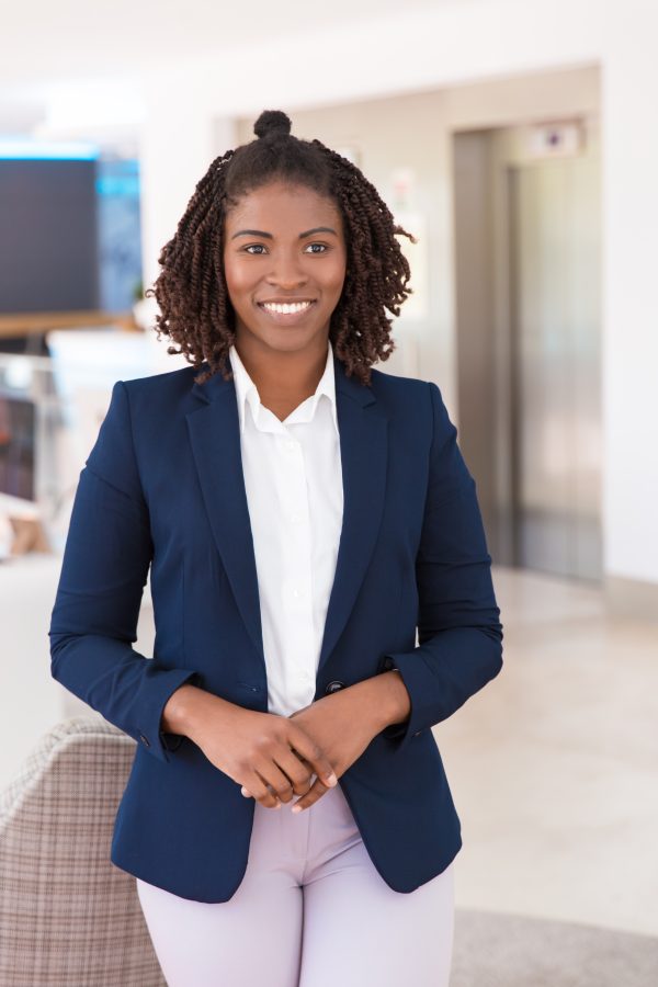 Happy young female professional posing in office hallway. Portrait of African American business woman in office suit standing for camera and smiling. Young business lady concept
